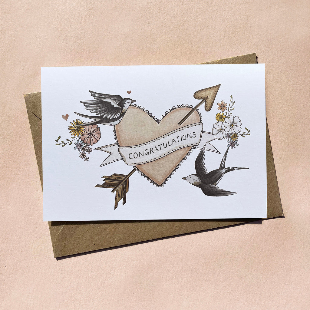 Greeting card with 'congratulations' banner over a vintage heart, cupid arrow, flowers and birds on an off white background. Blank inside.